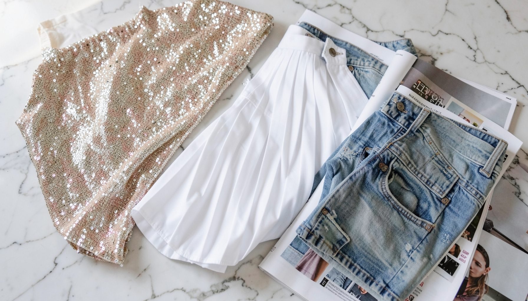 A mood board of fashion textures: shimmering sequin fabric, crisp white tennis pleats, faded vintage denim, and glossy magazine pages spread across a marble surface in soft natural light.