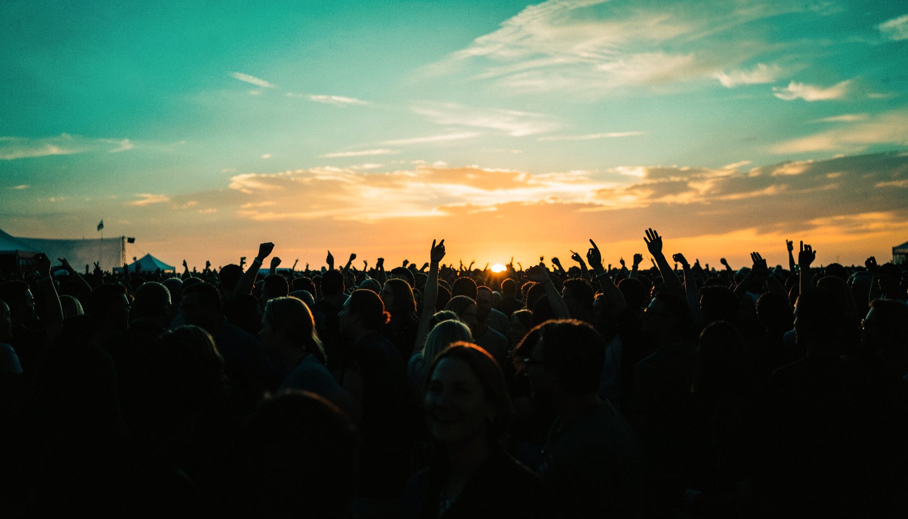 An outdoor music festival crowd silhouette against a warm sunset sky in teal and amber hues